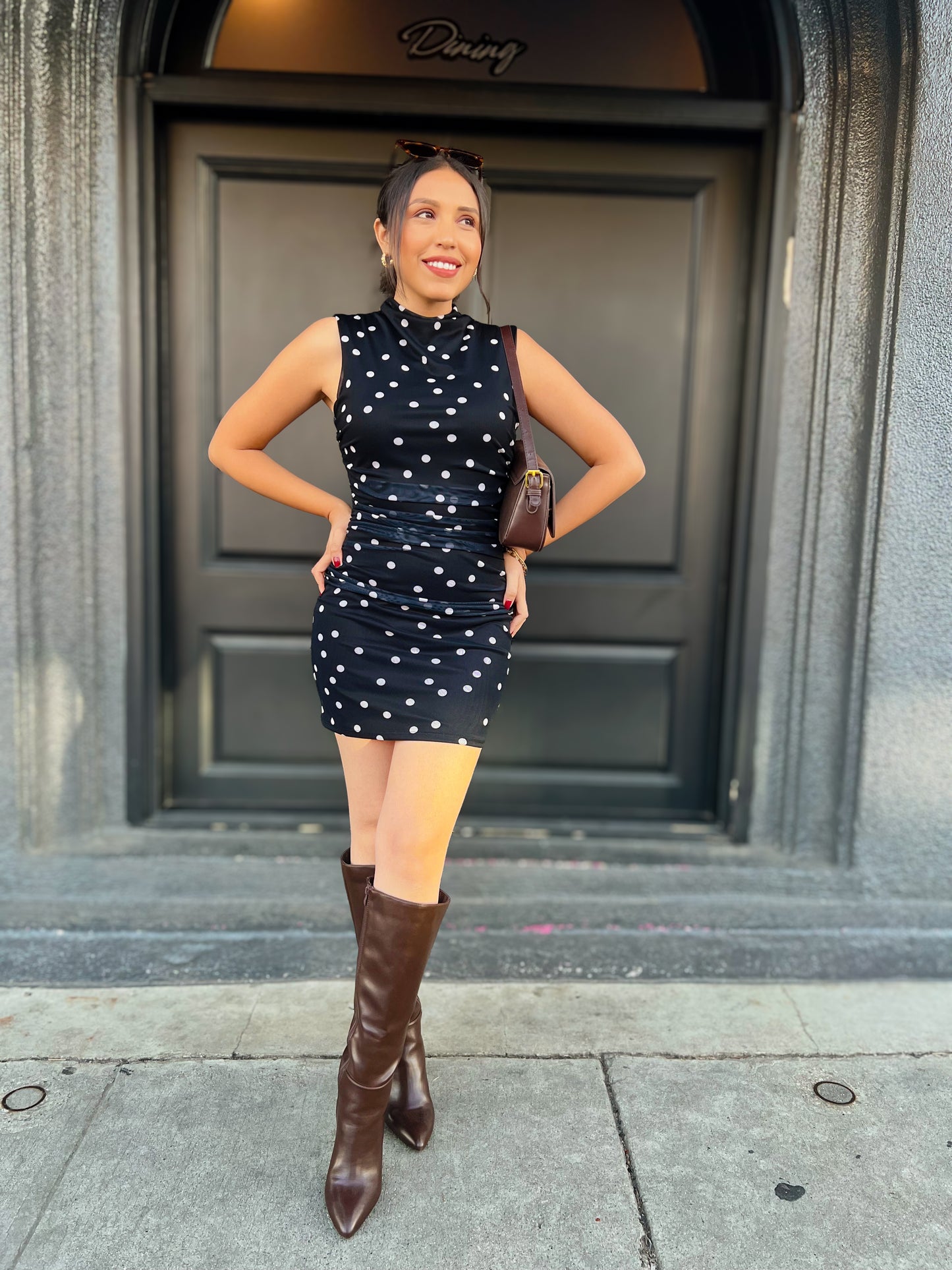 Woman in a black dress with white polka dots and brown knee-high boots standing in front of a building entrance.