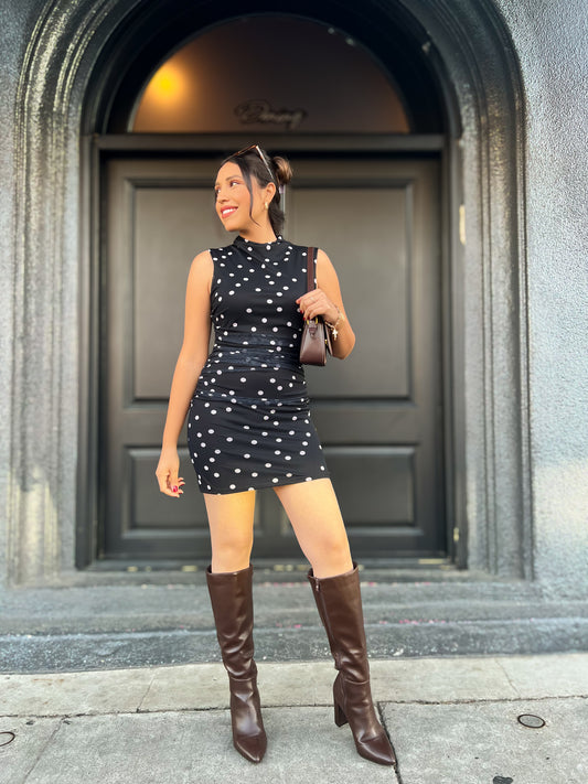 Woman in a black polka dot dress and brown knee-high boots standing in front of a building entrance.
