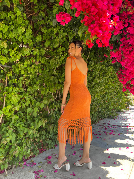 A brown skinned latina girl is wearing a body con bright orange crochet midi dress that has fringes at the hem. She is standing against a wall of bright magenta flowers that are blooming this spring. She is pairing the dress with nude slip on sandals and has her hair in a bun.