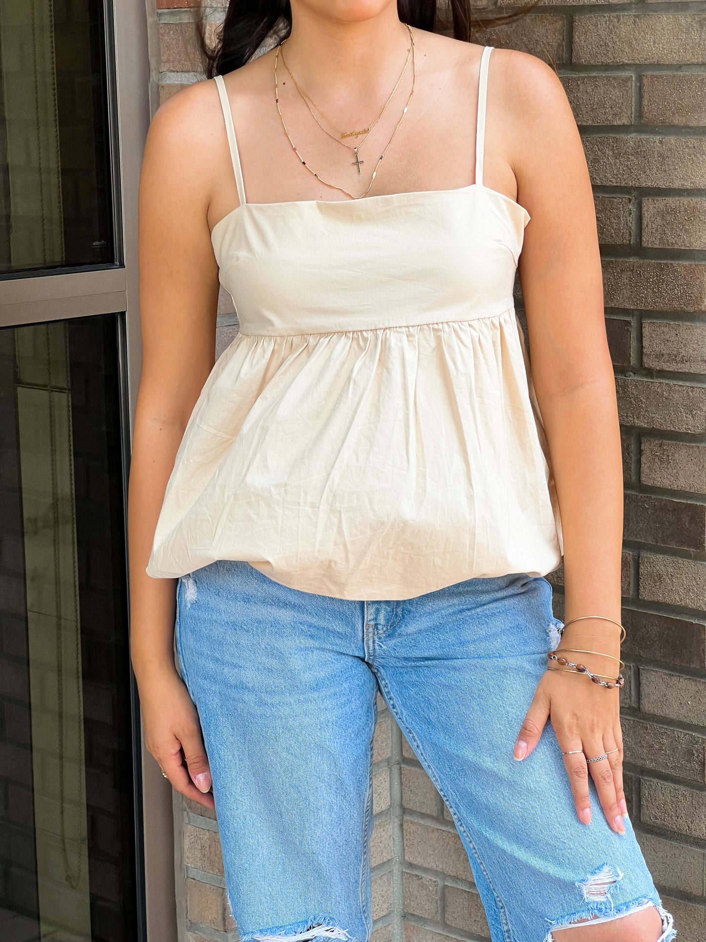 A close-up shop of a cream babydoll cami top. Accessorized with 3 golden necklaces and light wash jeans.