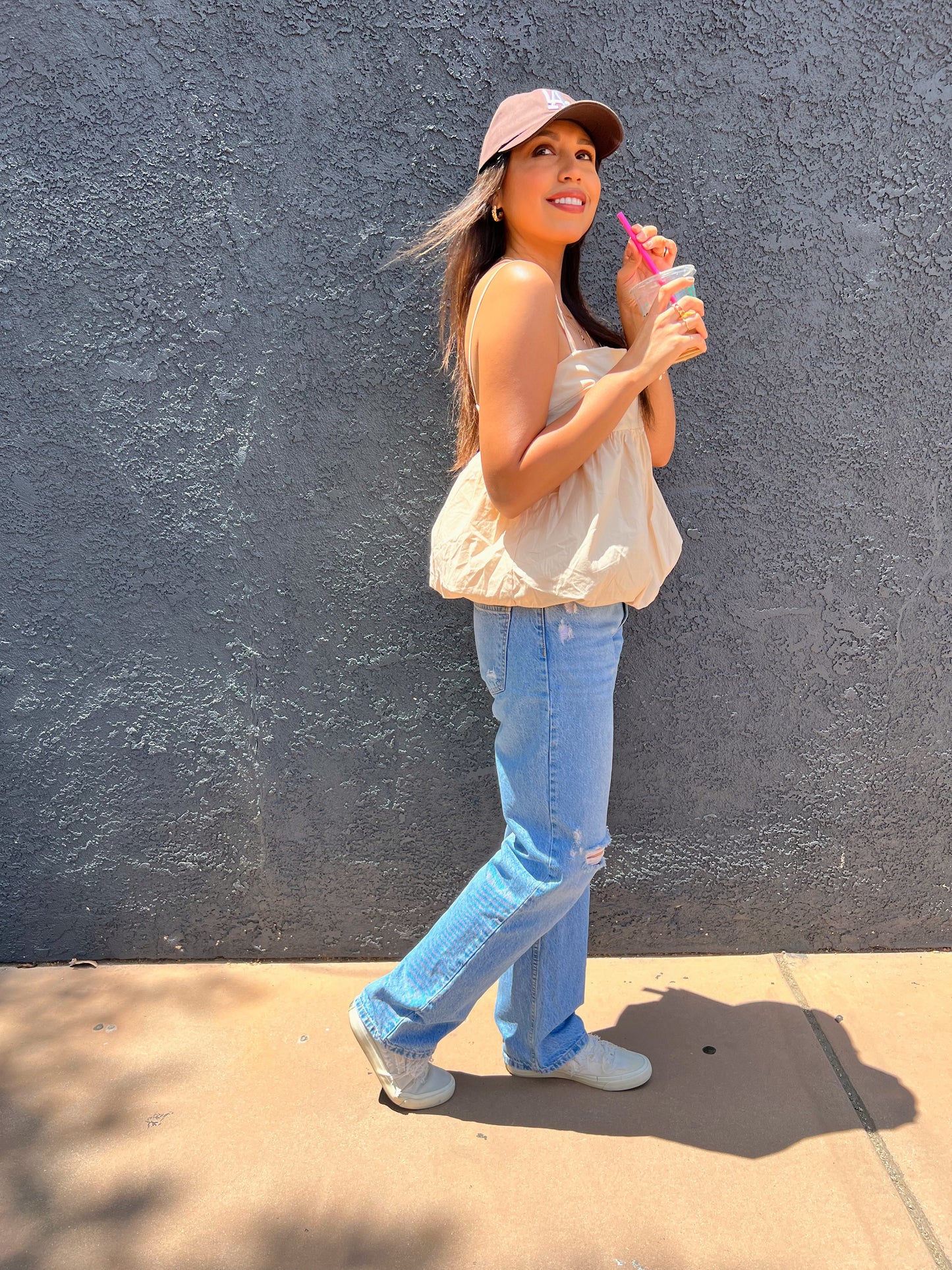 Woman smiling outdoors wearing a beige babydoll tank top, distressed light-wash jeans, and a maroon LA baseball cap, styled with gold layered necklaces and a brown shoulder bag and an iced coffee in her hand.