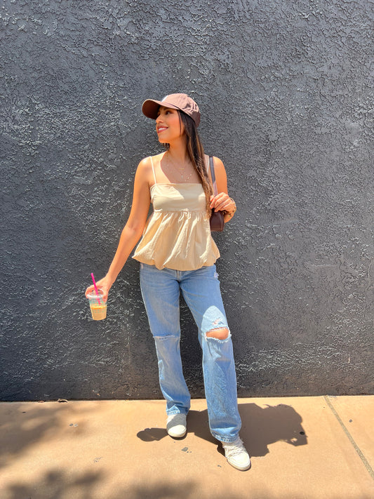 Model wearing a flowy beige babydoll summer top styled with light ripped jeans and a casual brown LA baseball cap, perfect for a sunny day look. While having an iced coffee in her hand.