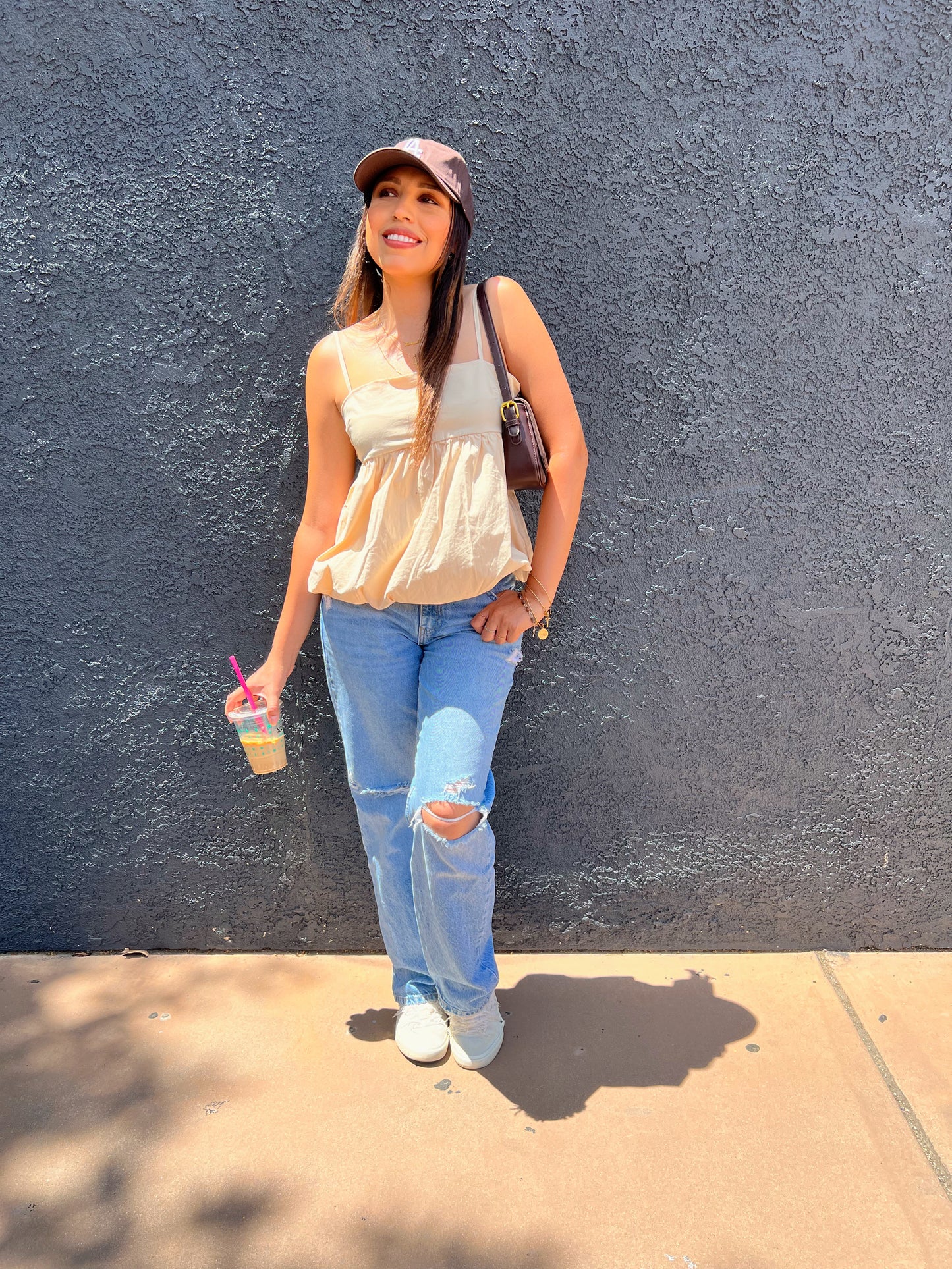 Woman smiling outdoors wearing a beige babydoll tank top, distressed light-wash jeans, and a maroon LA baseball cap, styled with gold layered necklaces and a brown shoulder bag.