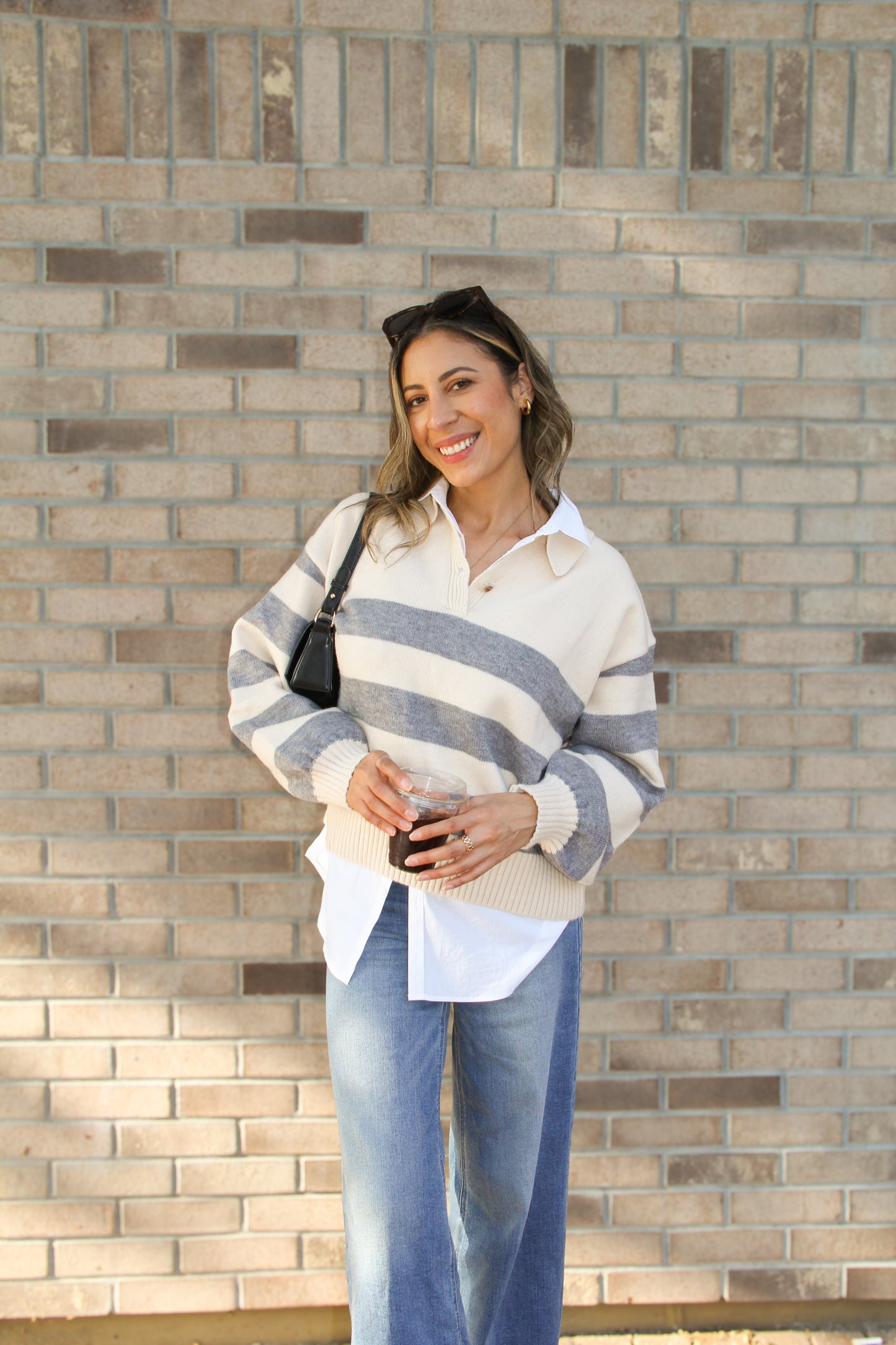 Woman wearing a striped sweater over a shirt with jeans, standing against a brick wall.