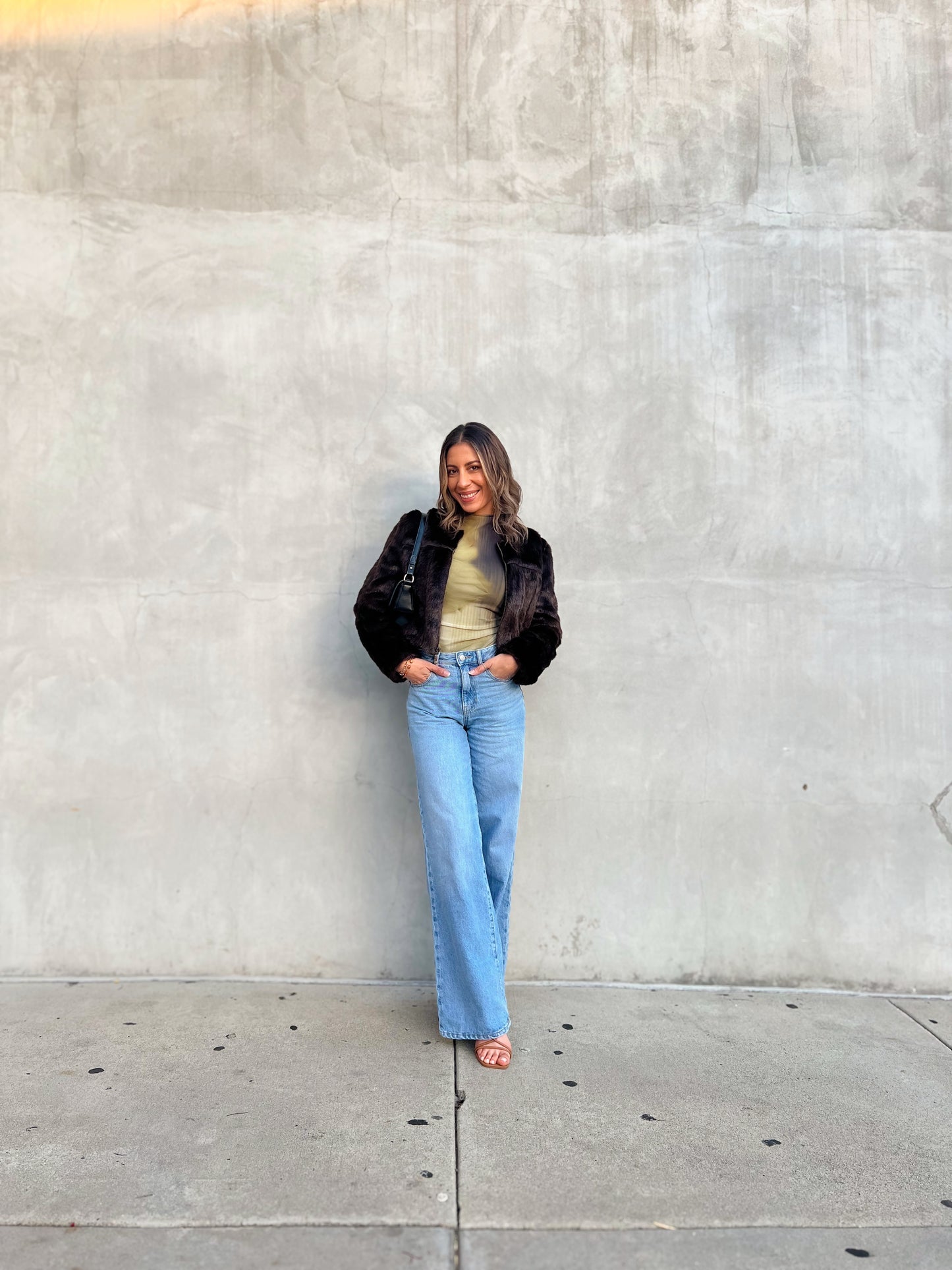 Person wearing a black jacket, beige top, and blue jeans standing against a gray wall.