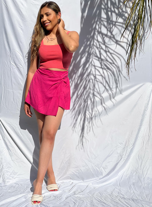 A brown-skinned latina model is wearing a corral ribbed knit crop top and pairing it with a hot pink skort. She has both hands on her waist. She styled this beautiful top with three golden necklaces. The chasing sunsets top has wide shoulder straps and has a square neckline. We also have this top available in off-white. The model is wearing a size small. This top and skirt will be the ultimate spring and summer piece because of it's vibrant colors!
