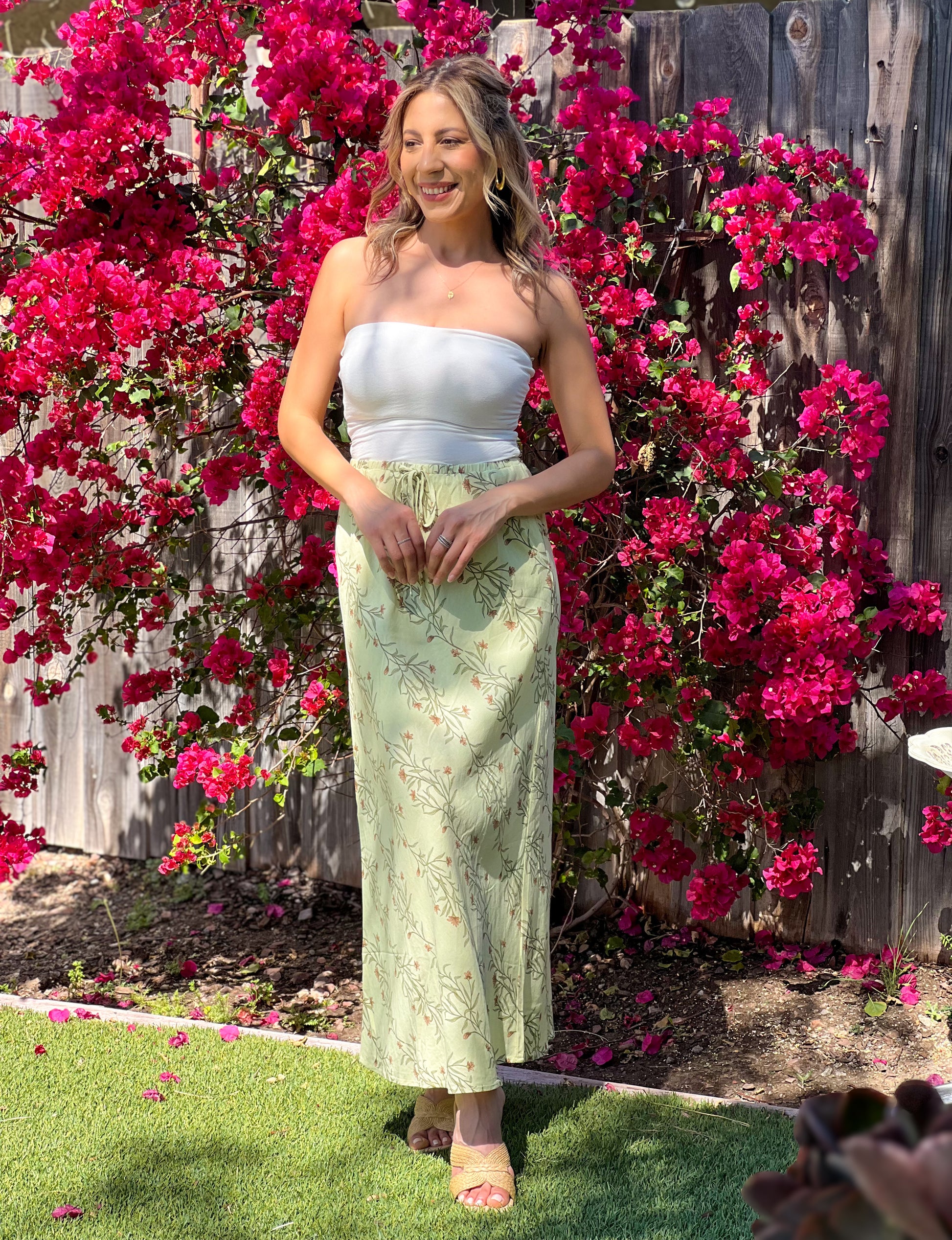 Woman in a strapless top and light green skirt standing in front of pink flowers.