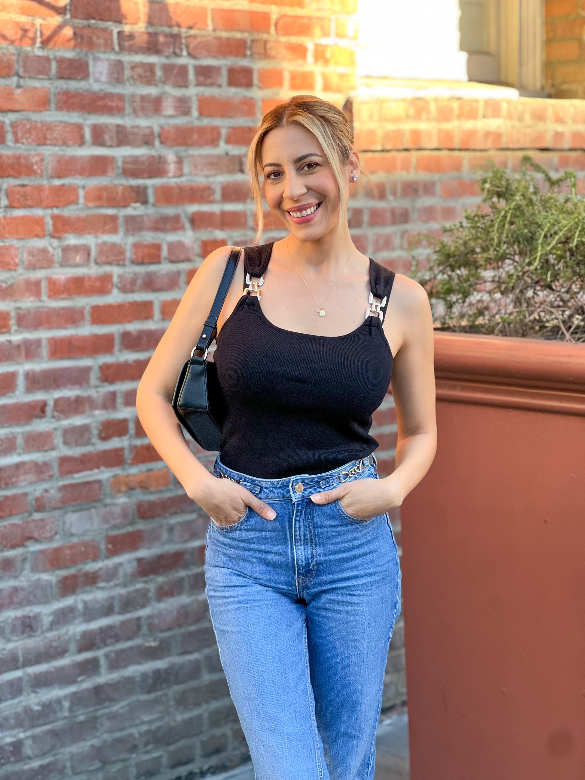 Woman in black tank top and blue jeans standing against a brick wall.
