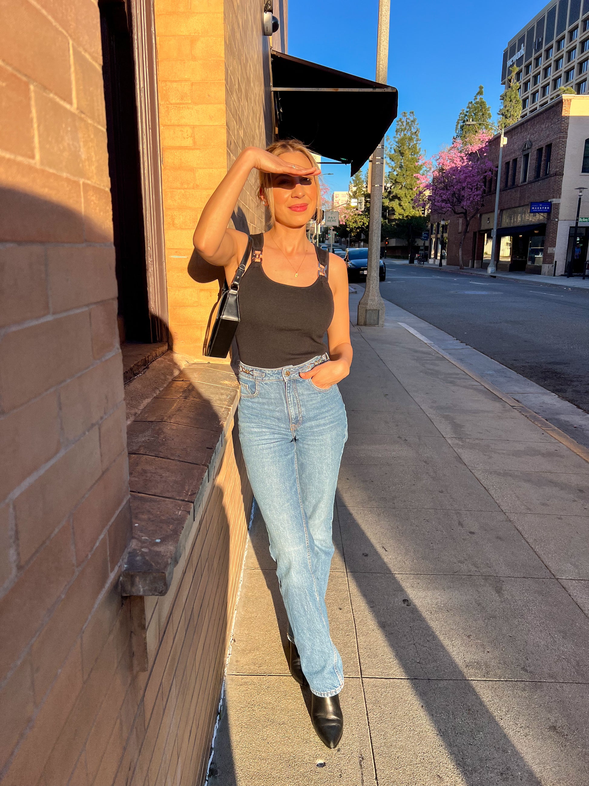 Woman standing on a city sidewalk wearing a black tank top and light blue jeans.