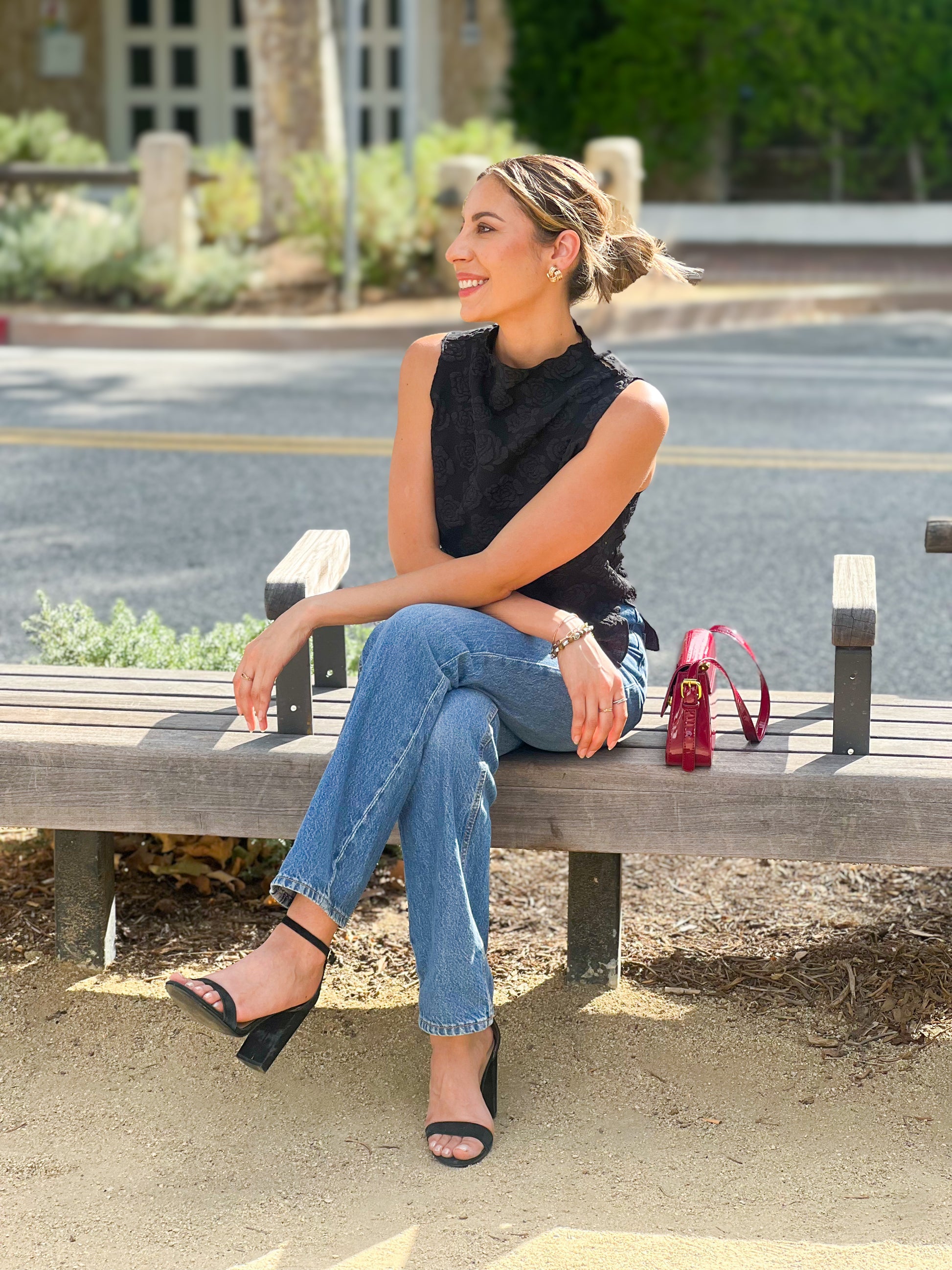 Woman sitting on a bench wearing a black sleeveless top and blue jeans, with a red handbag beside her.