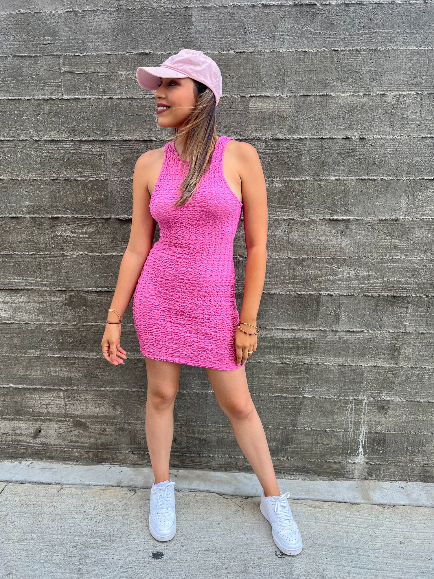 A brown skinned latina girl is standing in front of a white bricked wall. She is wearing a baby pink sleeveless short dress. She is pairing it with a pink baseball cap and white nike air force ones. Her accessory on her arm is a hot pink rectangle purse.