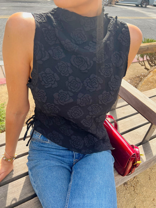 Person wearing a black sleeveless top with floral pattern and blue jeans, sitting on a bench with a red handbag.