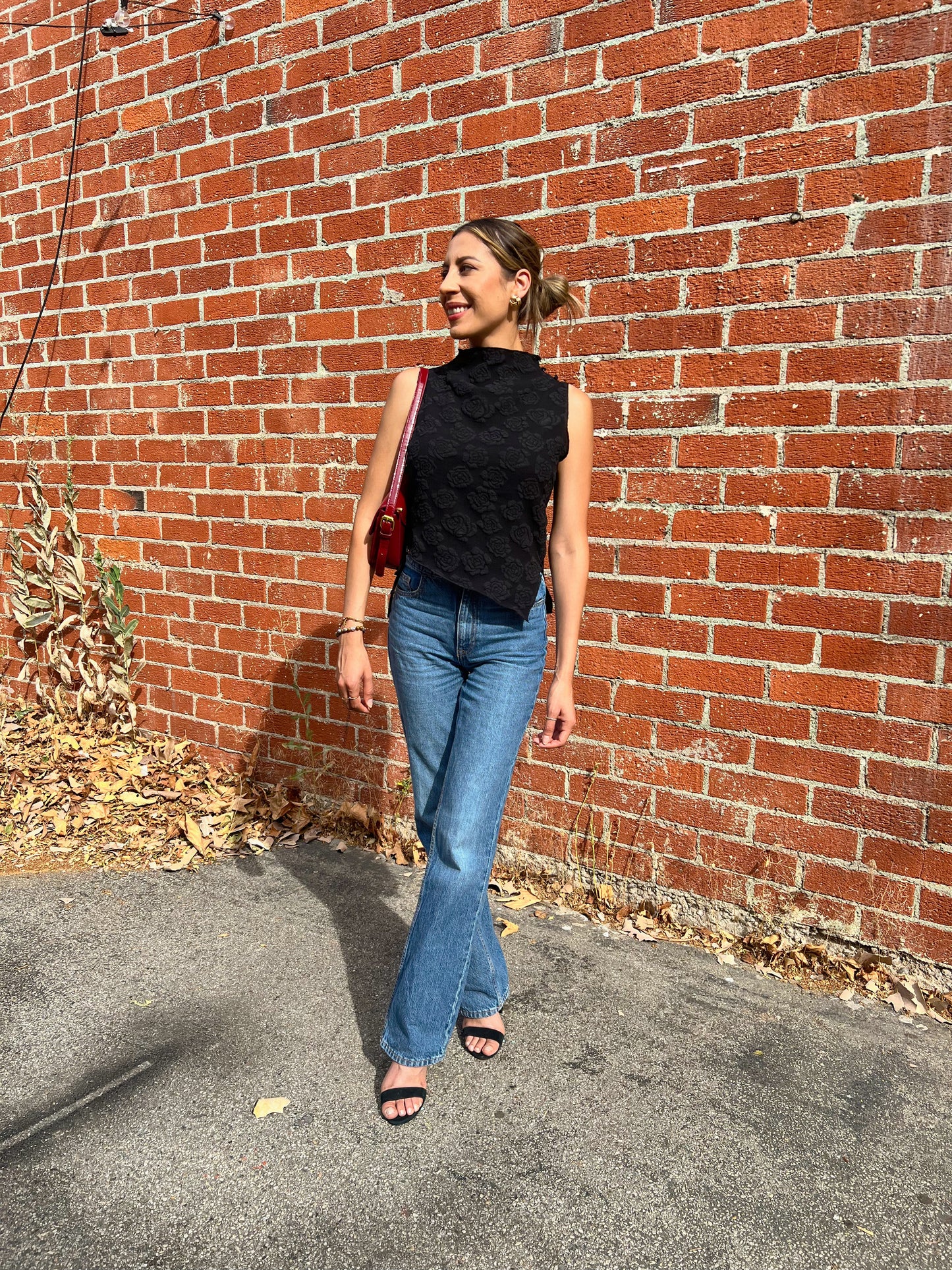 Woman in black sleeveless top and blue jeans standing against a brick wall.