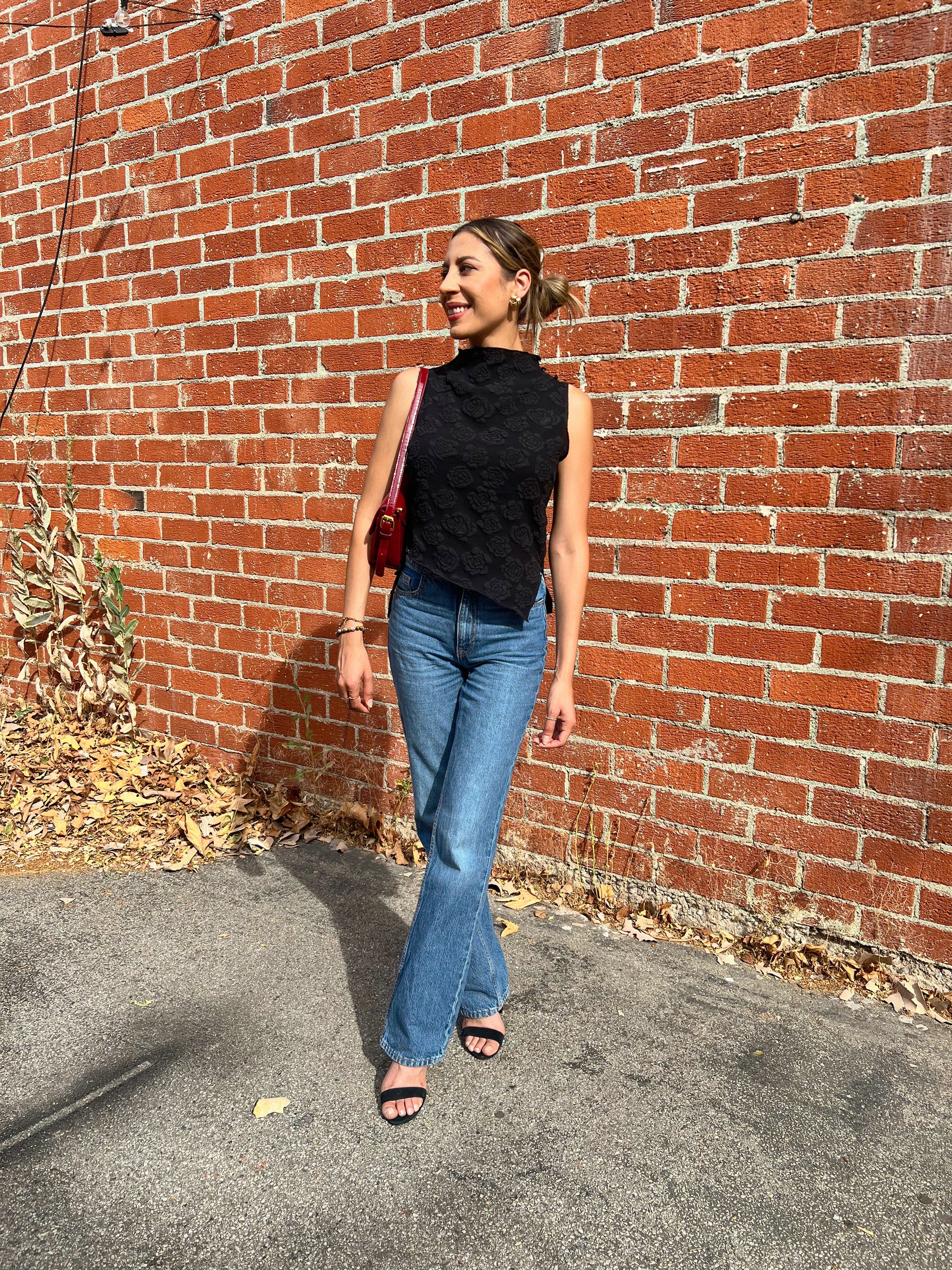 Woman in black sleeveless top and blue jeans standing against a brick wall.