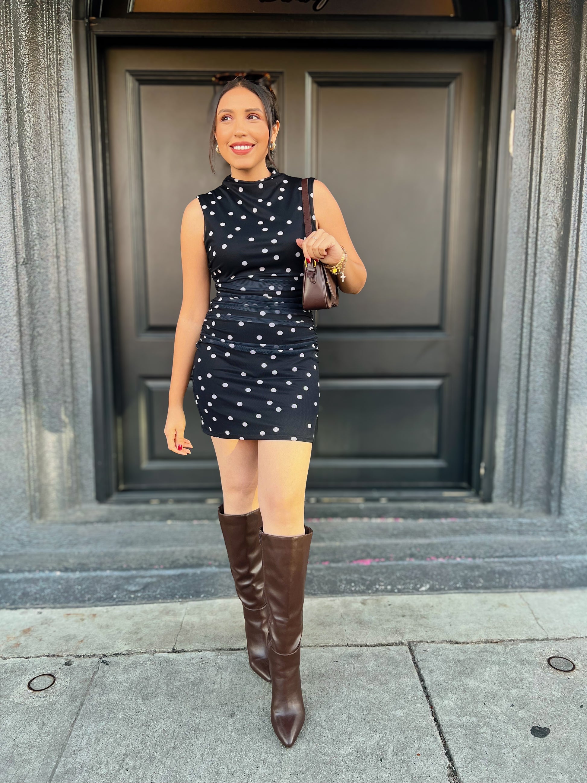 Woman in a black dress with white polka dots and brown knee-high boots standing in front of a building entrance.