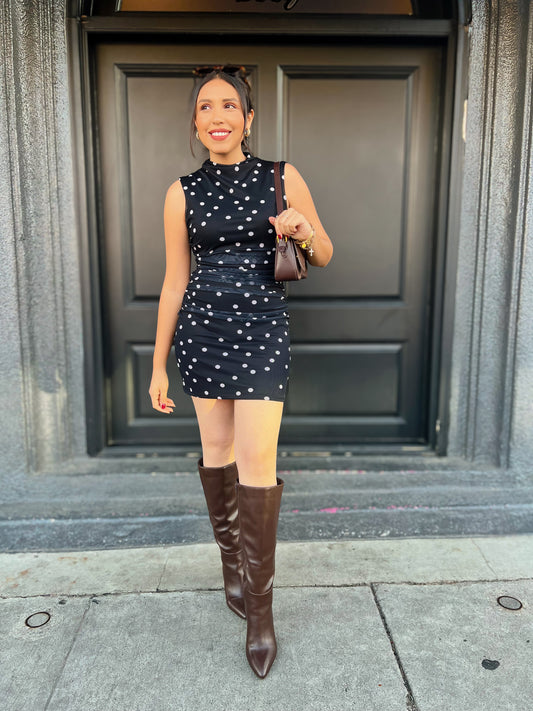 Woman in a black dress with white polka dots and brown knee-high boots standing in front of a building entrance.