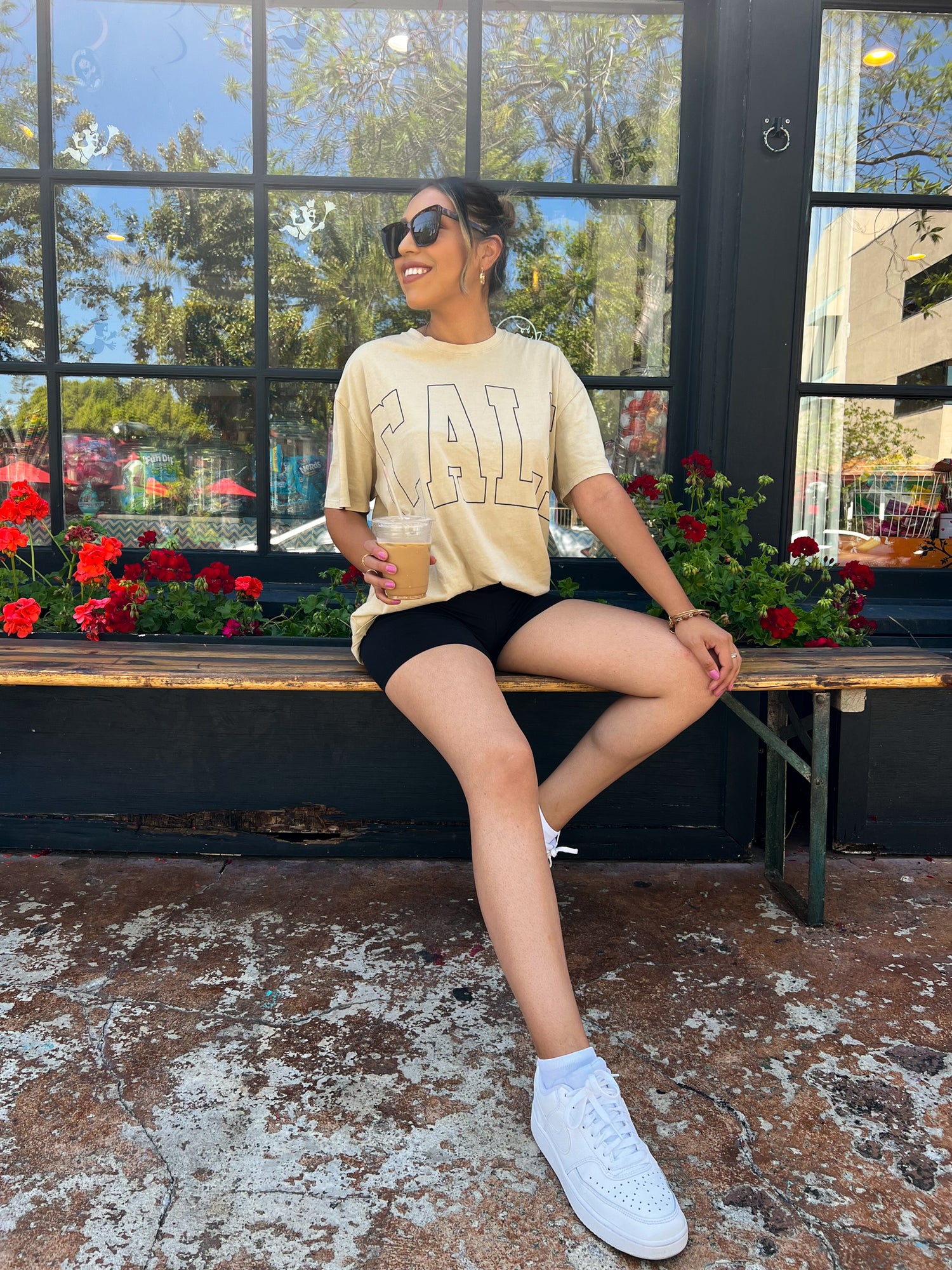 A brown skinned latina girl is wearing black short biker shorts and an oversized beige t-shirt. She's pairing it with white air forces and cat eye sunglasses. She has a vanilla latte on her right hand sipping on it. She is standing by a candy shop that has a black exterior and red bright flowers.