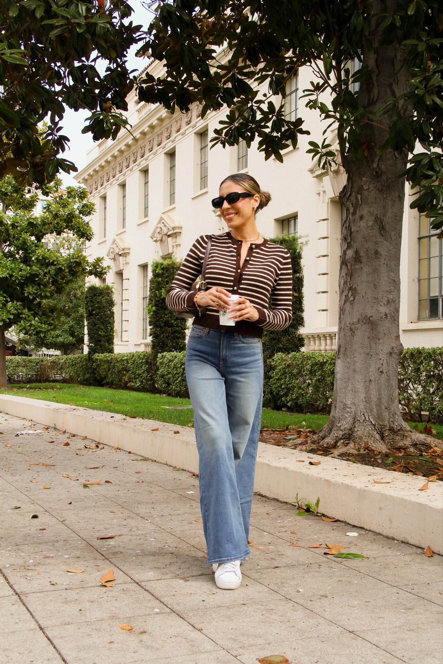 Woman walking on a sidewalk in an urban setting with trees and buildings in the background.