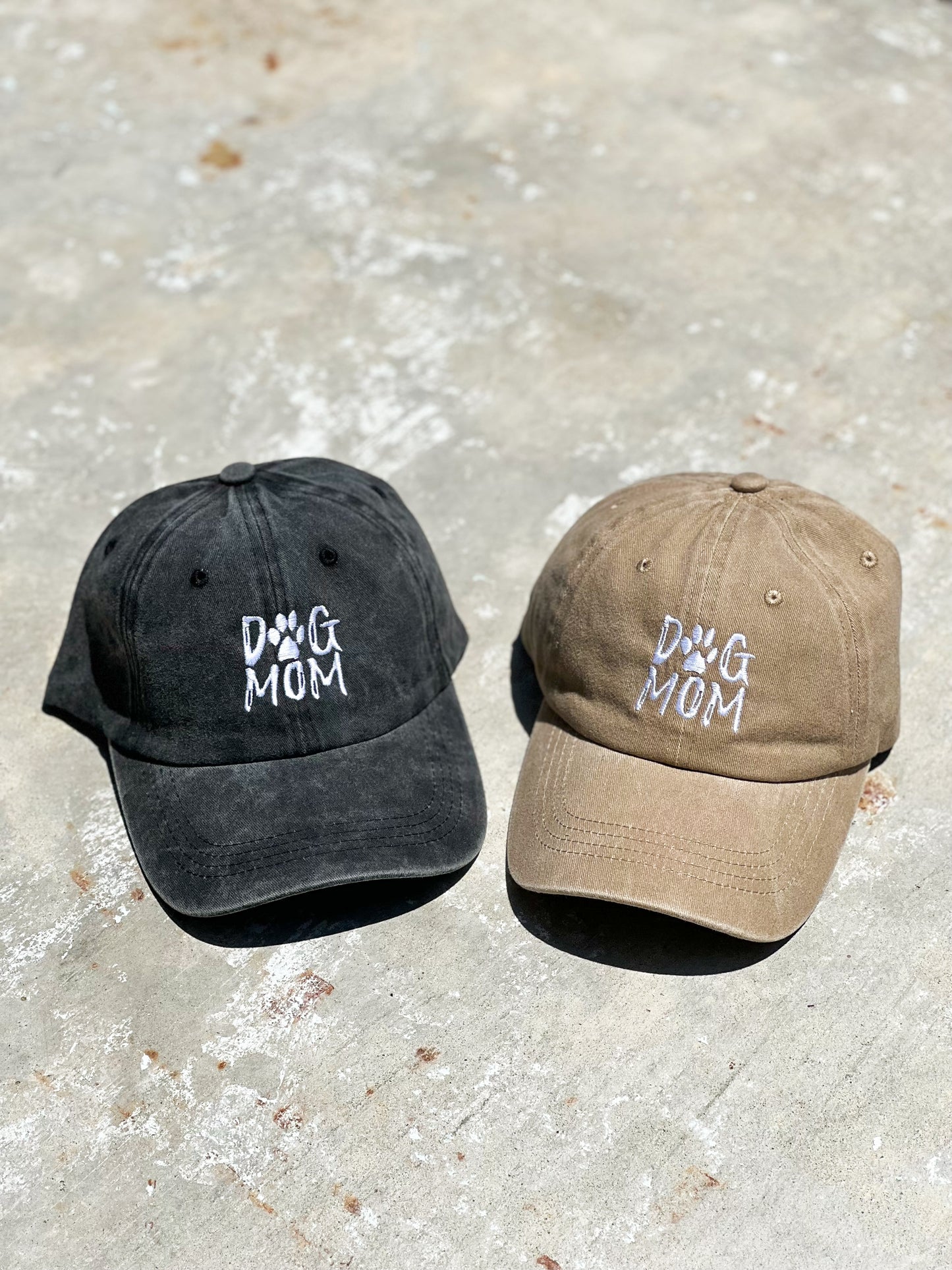 There is two baseball caps on a concrete gray floor. One baseball cap is charcoal black and has "dog mom" printed on the front. Next to it is a beige nude baseball cap with a "Dog Mom" print on it in the front.