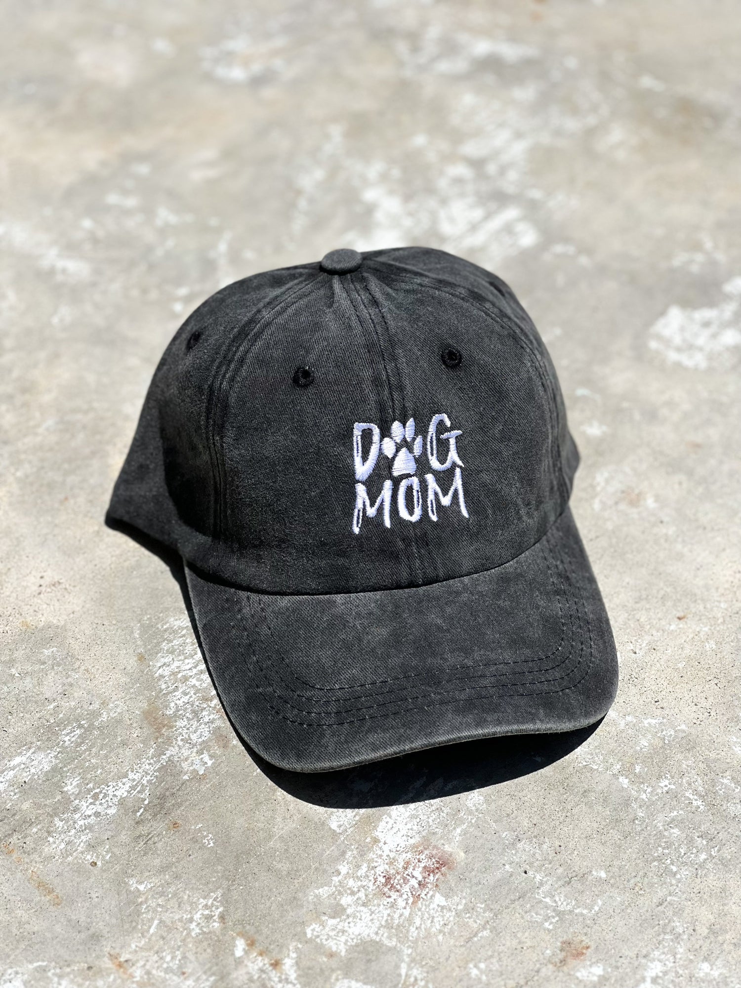 There's a black baseball distressed cotton cap with "Dog Mom" printed on the front of the cap. It's laying on the gray concrete floor.