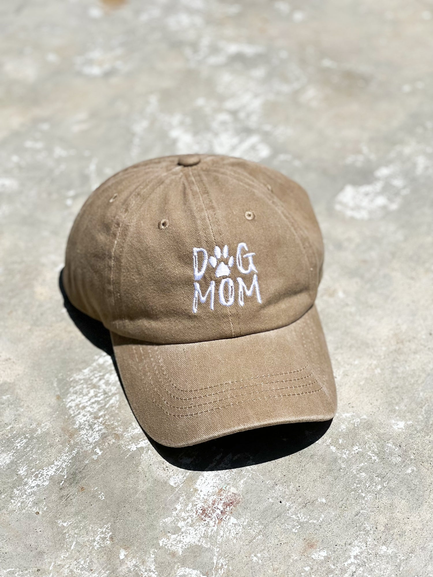 There's a beige baseball distressed cotton cap with "Dog Mom" printed on the front of the cap. It's laying on the gray concrete floor.