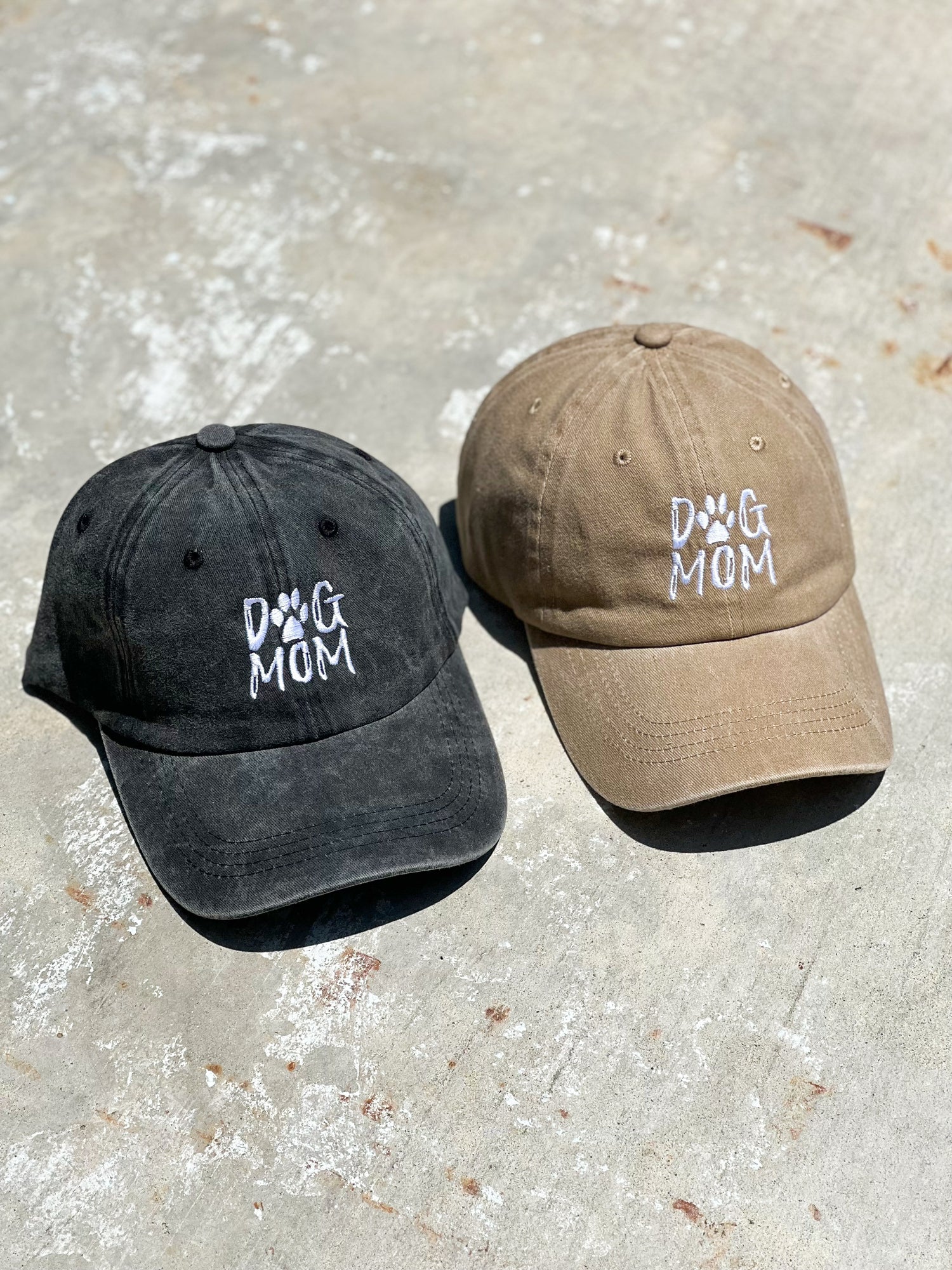 There is two baseball caps on a concrete gray floor. One baseball cap is charcoal black and has "dog mom" printed on the front. Next to it is a beige nude baseball cap with a "Dog Mom" print on it in the front. 