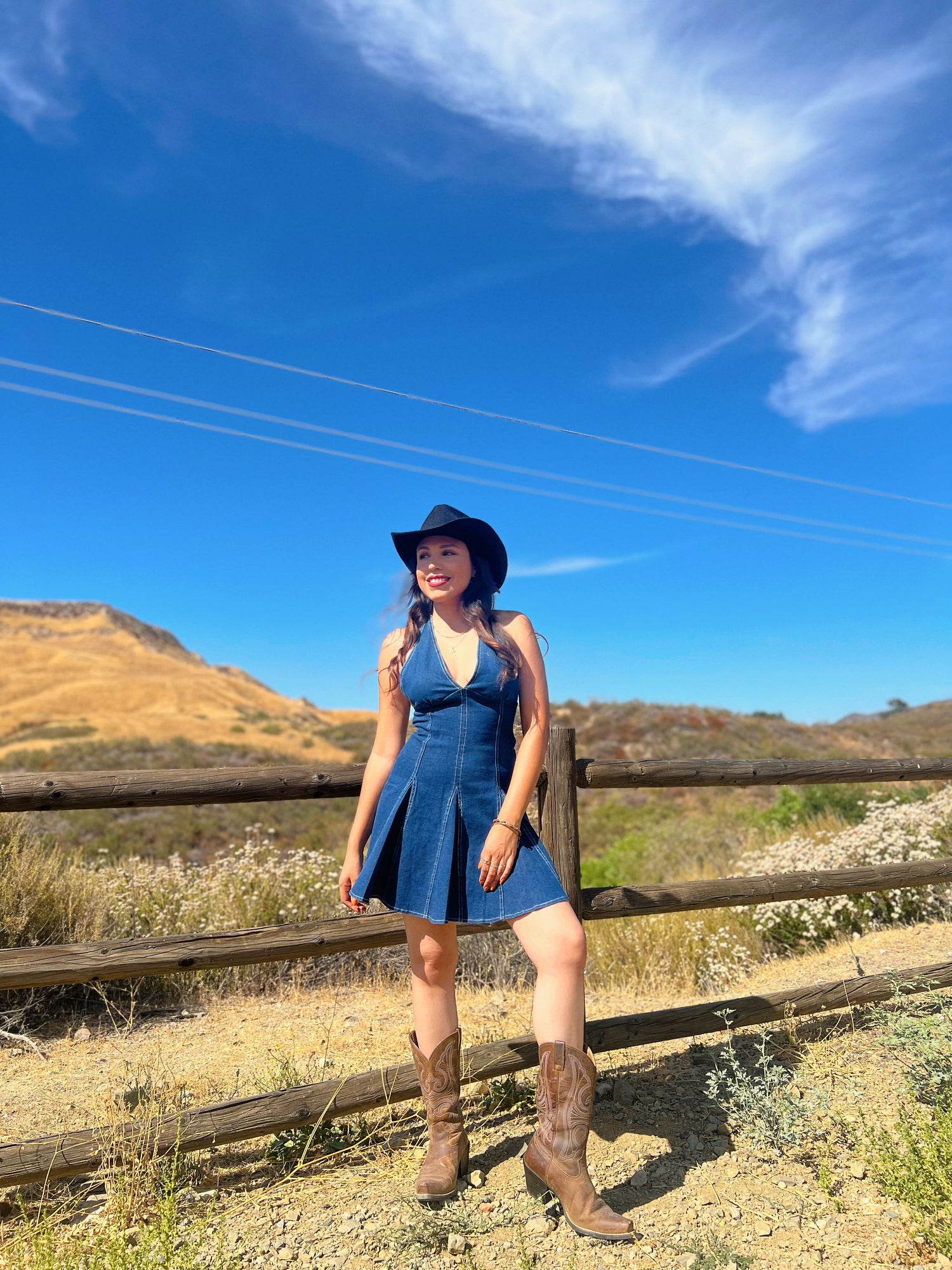 Woman in a blue dress and cowboy boots standing in a field with a wooden fence and clear sky.
