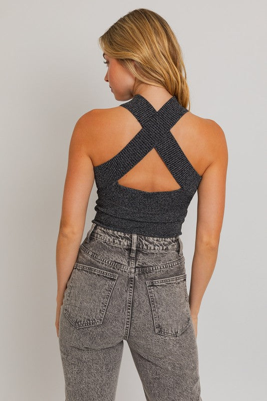 This is back view of a model showing the backside of the metallic bodysuit. We see that there’s a criss cross shaped. This top is sleeveless. She is wearing gray denim jeans. Both of her arms on the side. 