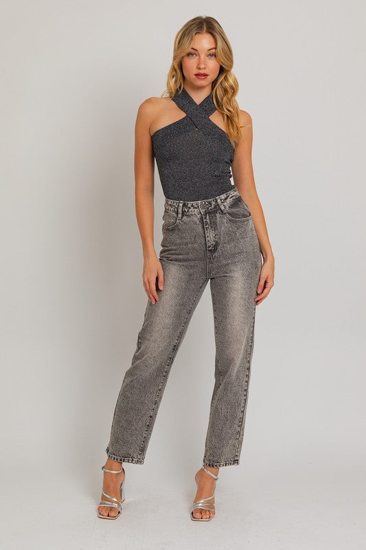 This is a full body picture of a Caucasian blonde woman posing wearing a metallic black bodysuit and gray denim jeans. Her heels are metallic silver and open toed. Her long hair is placed in front of her and has both arms beside her. 