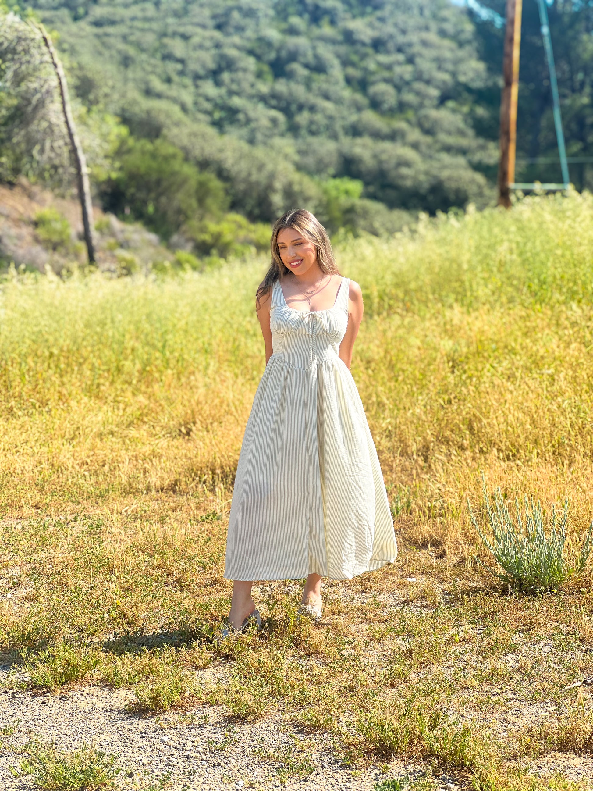 Woman in a white dress standing in a natural setting with greenery and a dirt path.