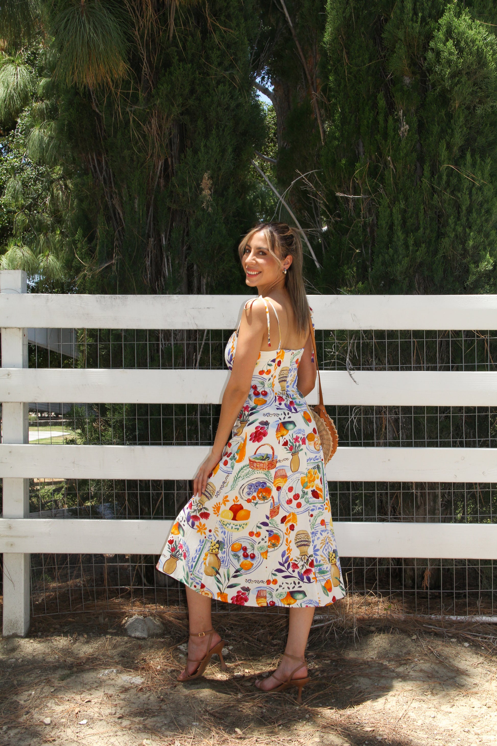 Woman in a floral dress standing in front of a white fence with greenery in the background