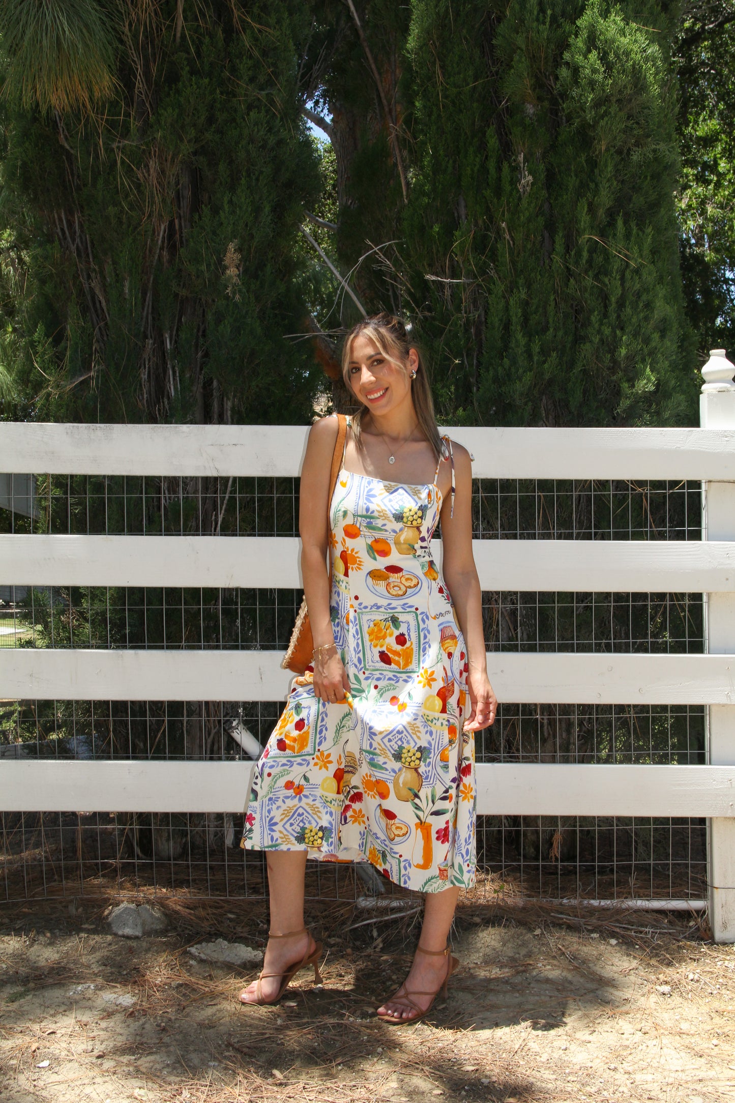 Woman in a floral dress standing in front of a white fence with greenery.