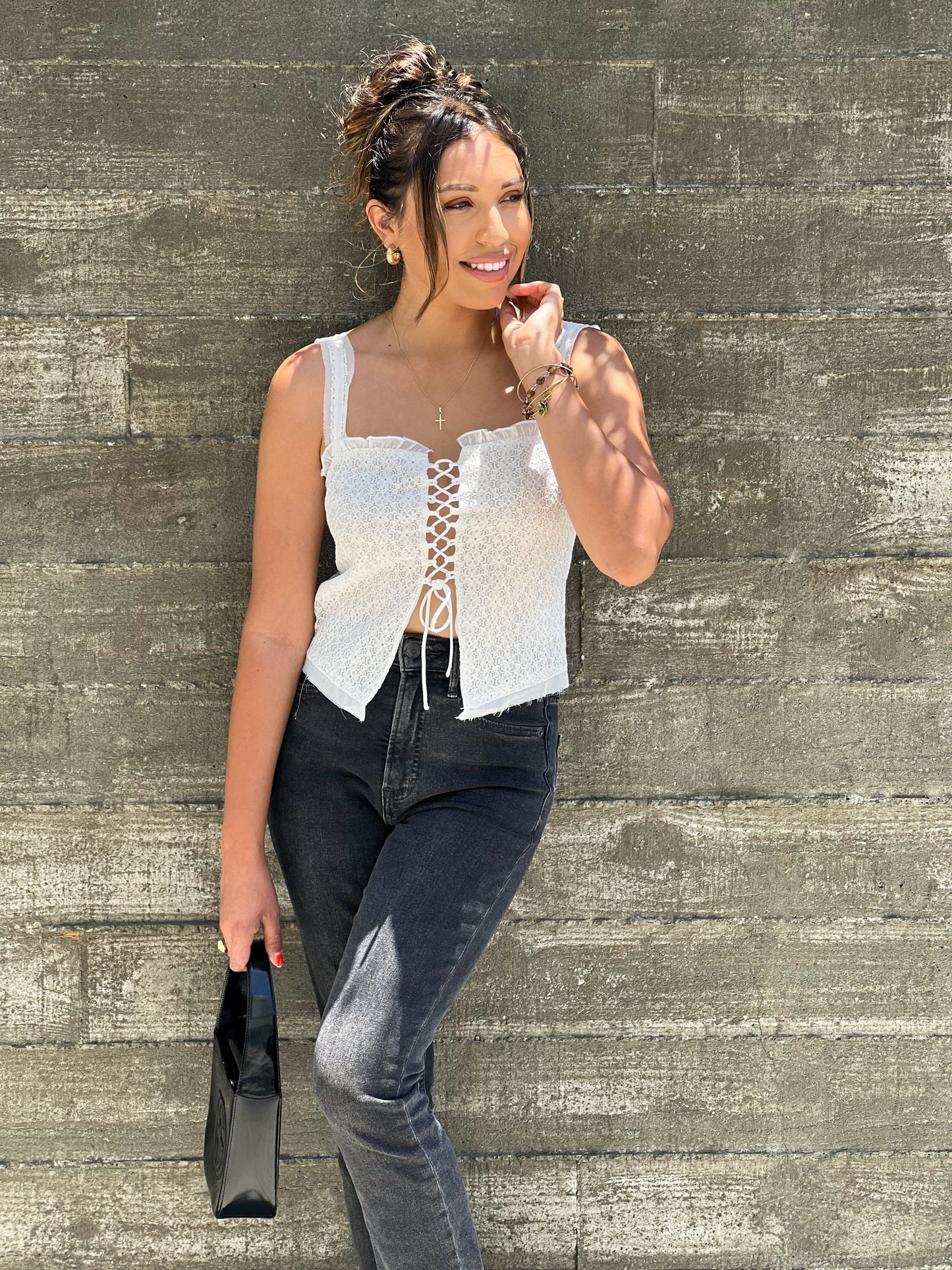 The model is tanned skinned and looking off to the side with one hand holding her hair. The other hand she is holding a black small handbag. She is wearing a white sleveless lace top and black jeans. 