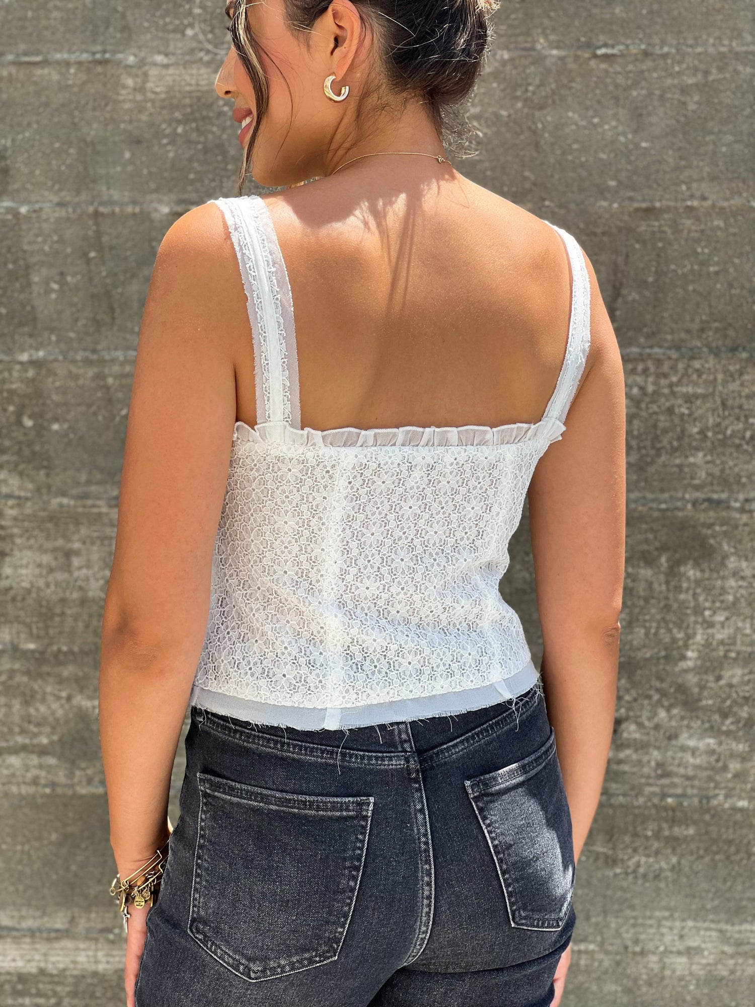 This is a closeup shot of the back of the shirt. The model is tanned skinned and wearing a white sleeveless lace top. It has a square neckline. She is wearing black jeans and has her hair in a bun. 