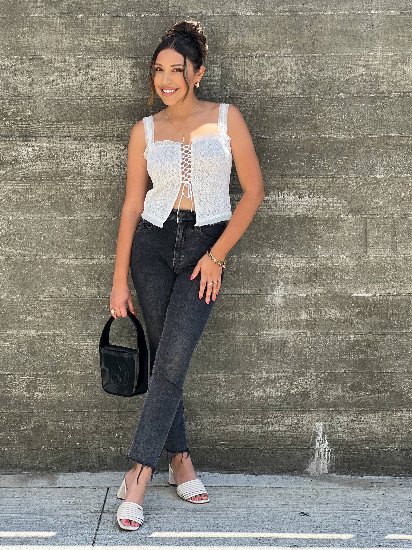 A brown skinned latina girl is wearing a white sleeveless lace string top. She paired it with black jeans and white sandals. She has her hair up on a bun to show the sweetheart neckline. She has gold bracelets on her right hand.