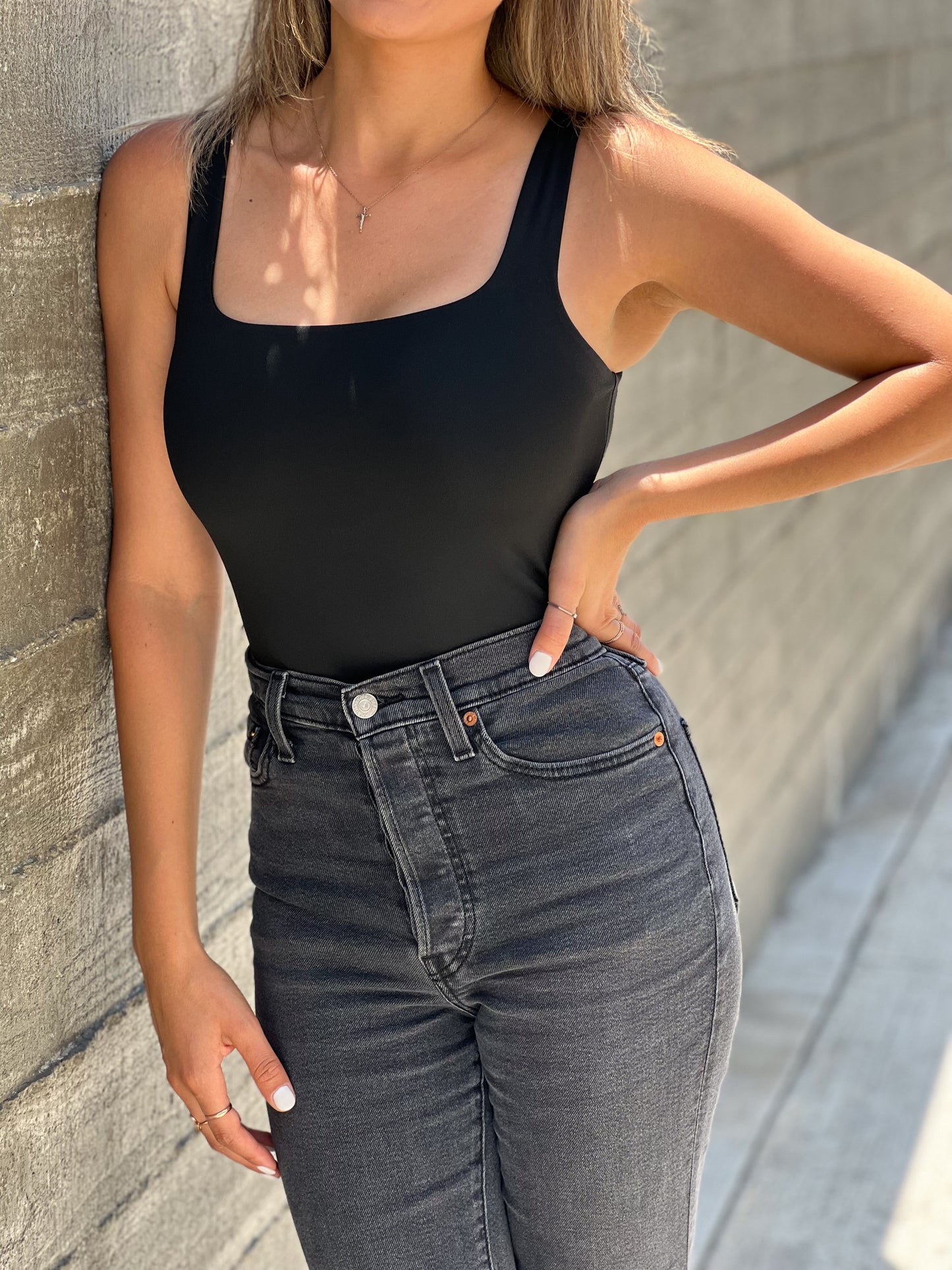 A brown skinned latina model is wearing a black bodysuit that has a squared neckline. She has one arm on her waist and the other touching her pants. Its a close-up shot of the top. Her face isnt showing.