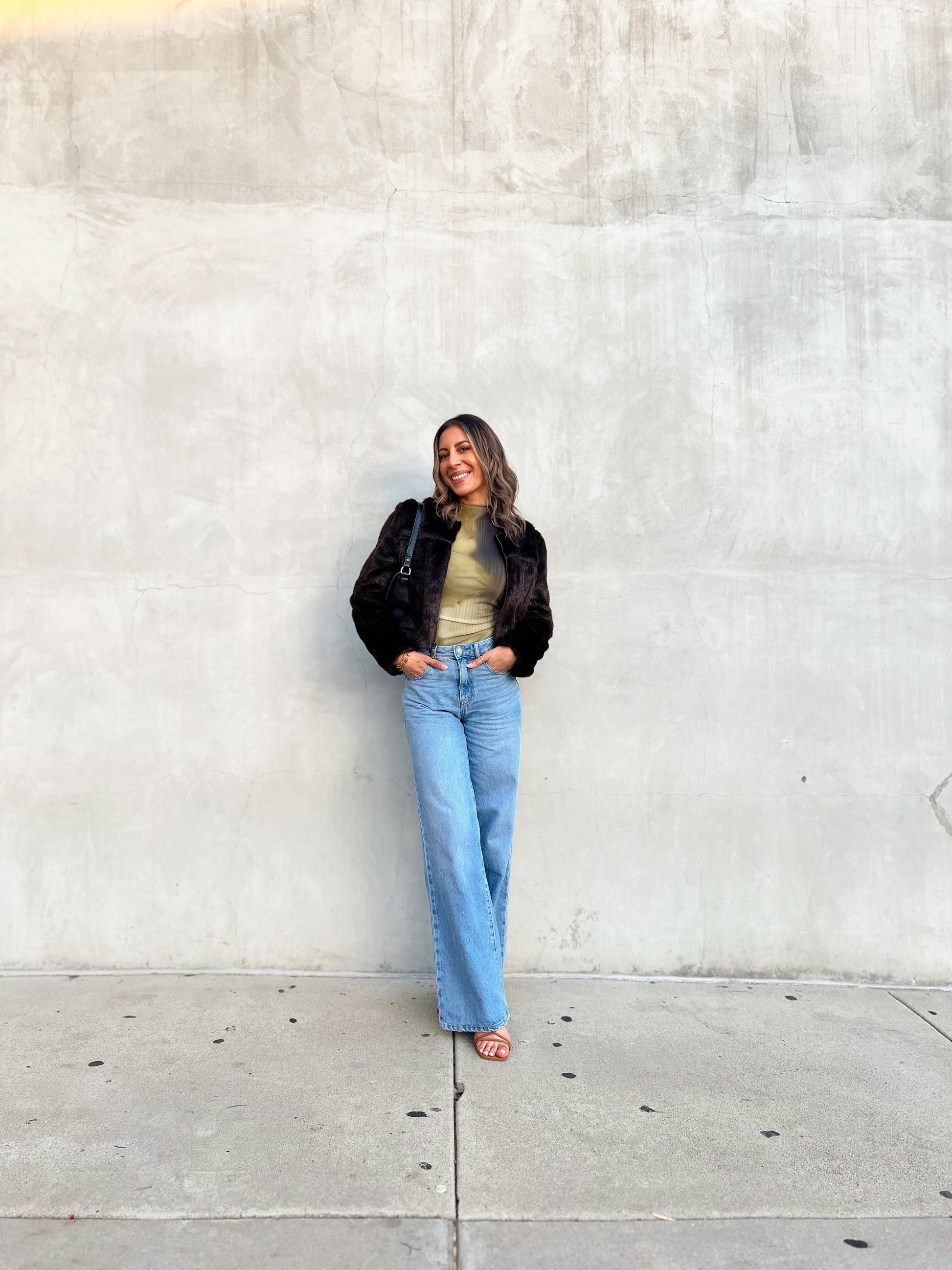 Woman standing against a light gray wall wearing a black jacket, green top, and blue jeans.