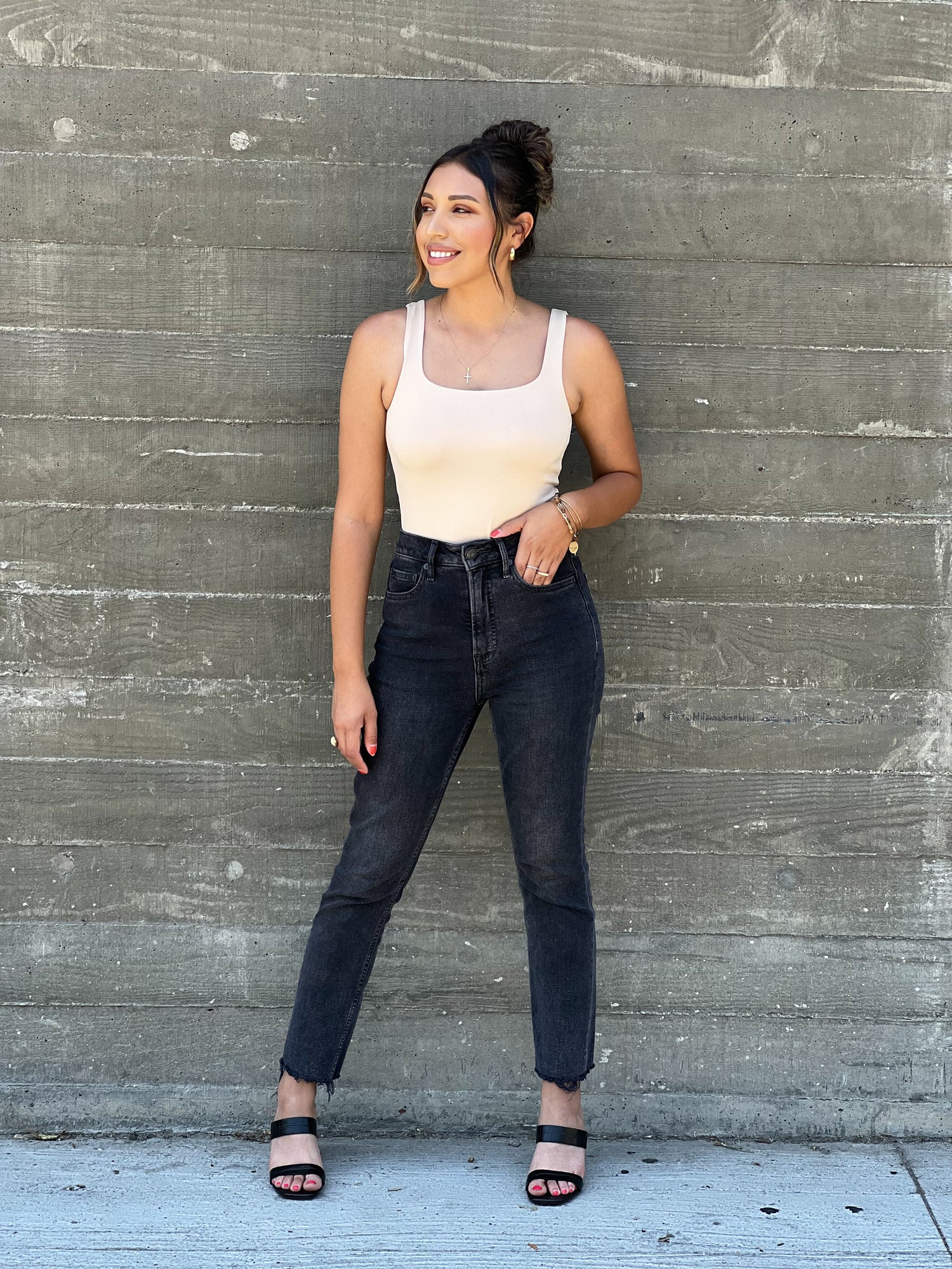A brown skinned latina model is wearing black jeans and a nude bodysuit. The bodysuit has a sqaure neckline. She has her hair up in a bun and wearing brown open toed heels. She has her right hand in the jeans pocket and the other one straight ahead. 