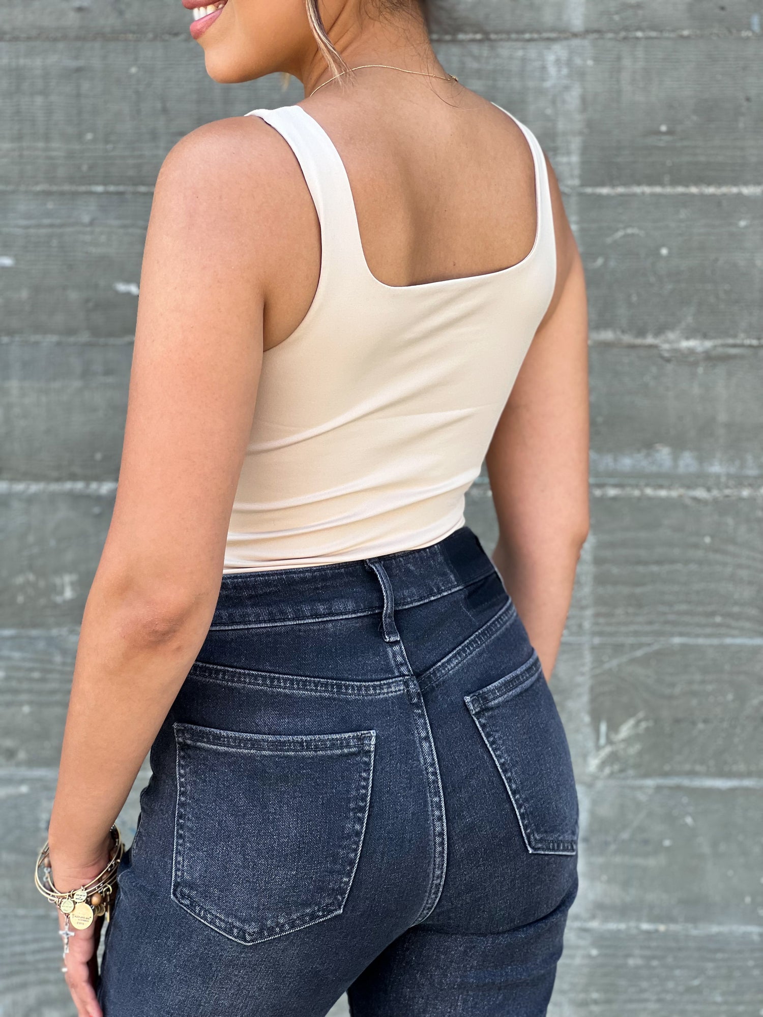 This is a closeup shot of the nude bodysuit. She is facing her back to the camera. The model is tan skinned and pairing the top with black jeans. 