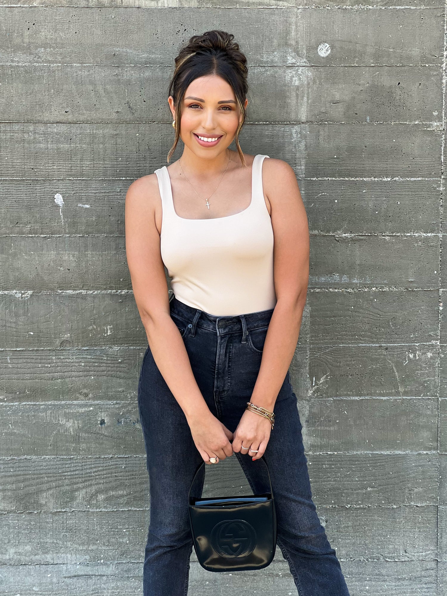 A brown skinned latina girl wearing a nude bodysuit with black jeans. She has her hair up in a messy bun and two front hairs in front of her face. She accessorized the look with a black leather gucci bag and gold hoop earrings and a gold cross necklace. She is posing with both arms facing toward her holding the bag.