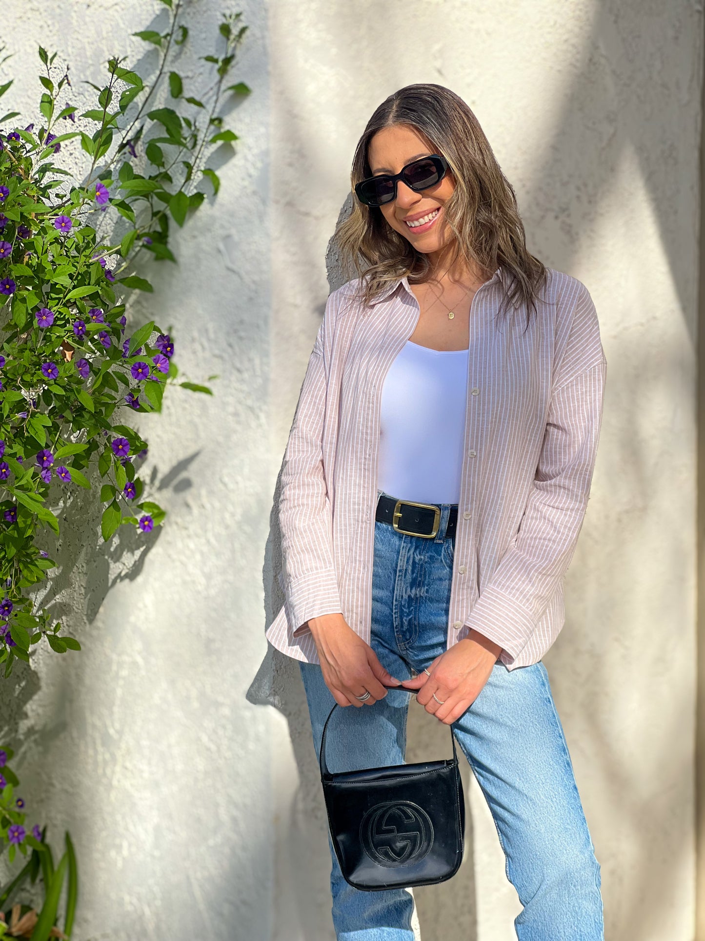 Woman standing outdoors against a white wall with greenery, wearing a light pink cardigan, blue jeans, and sunglasses.