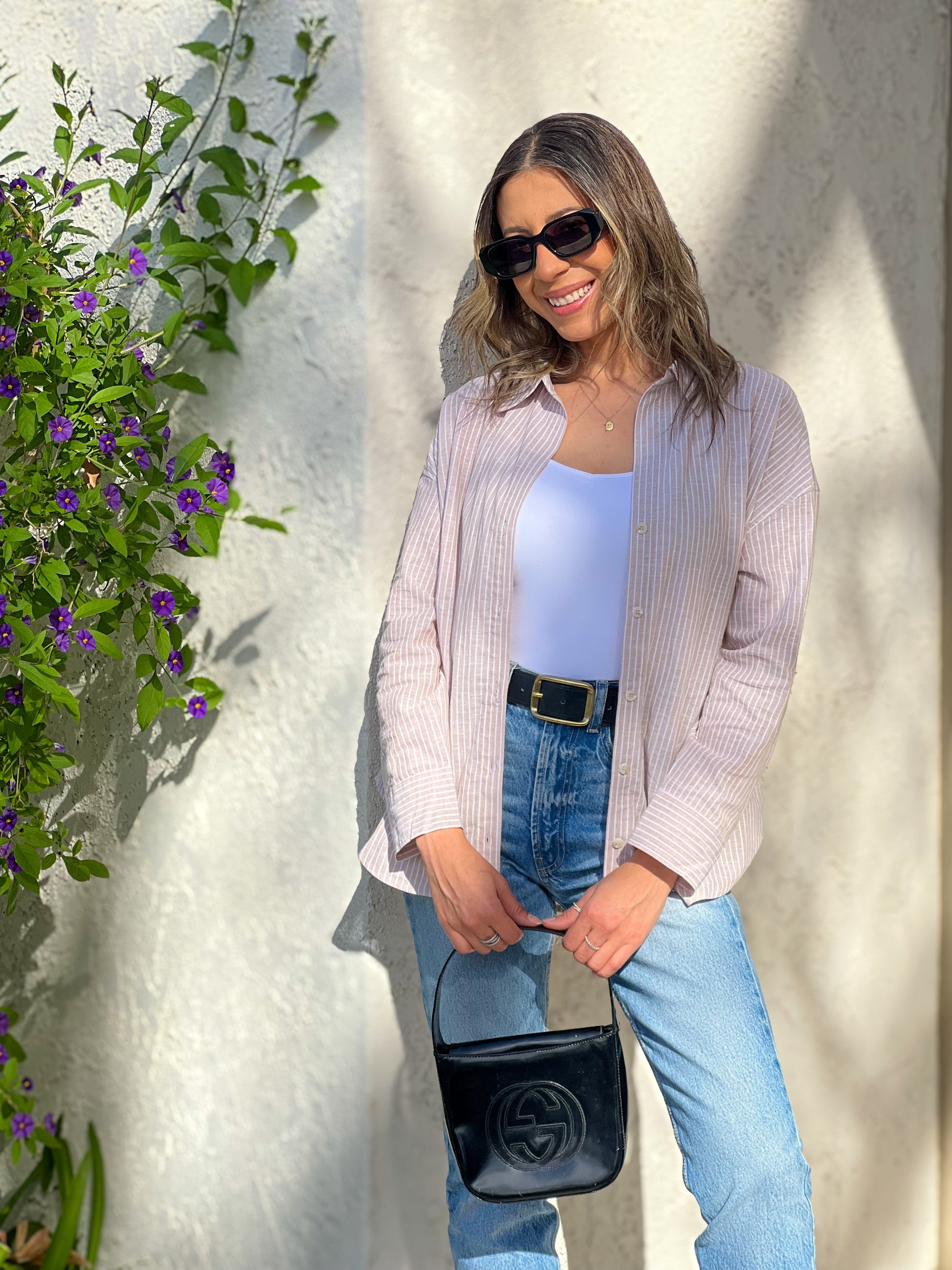 Woman standing outdoors against a white wall with greenery, wearing a light pink cardigan, blue jeans, and sunglasses.