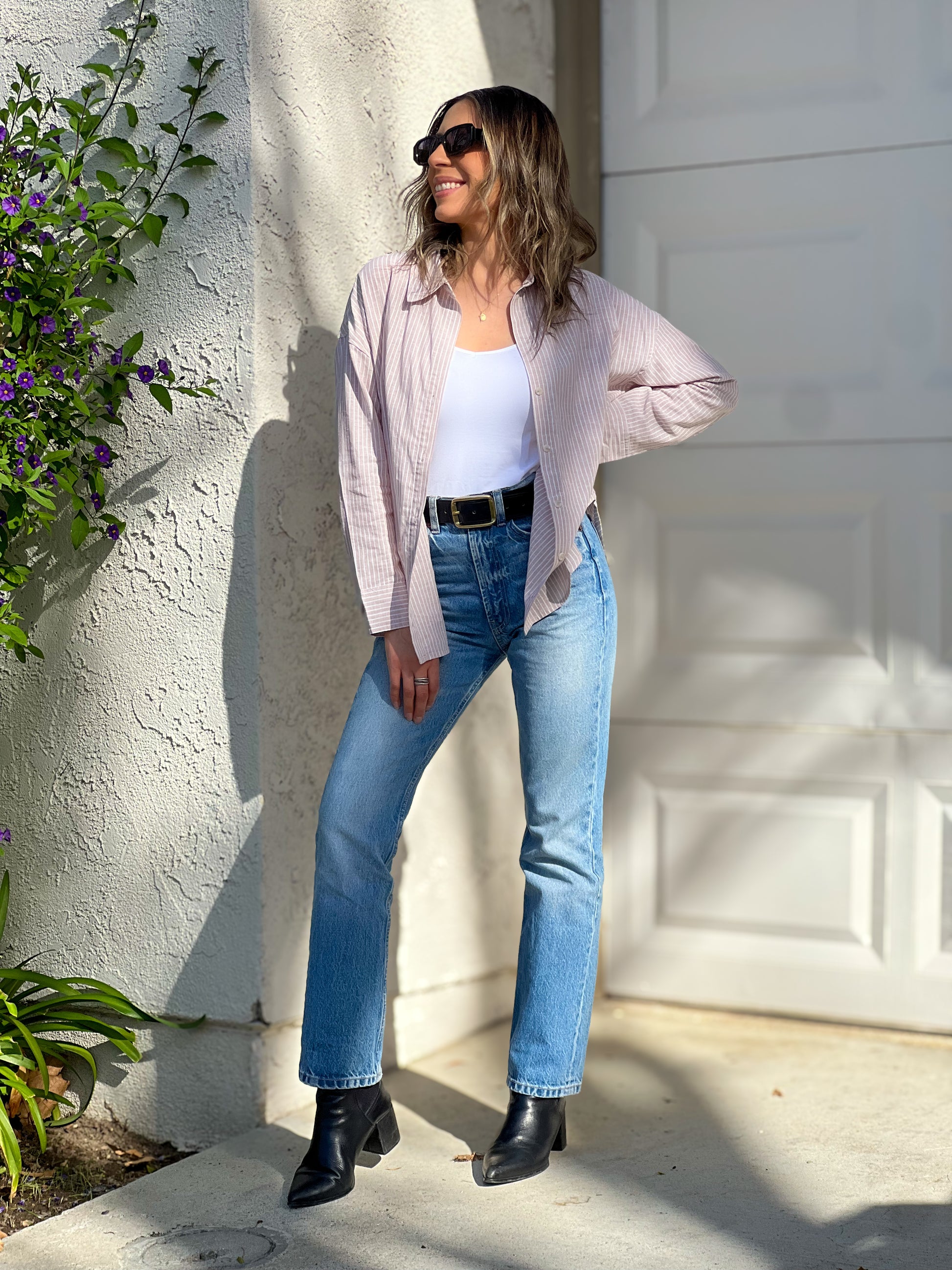 Woman standing against a white wall wearing a light pink shirt, white top, blue jeans, and black boots.