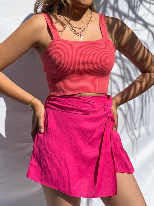 A brown-skinned latina model is wearing a corral ribbed knit crop top and pairing it with a hot pink skort. She has both hands on her waist. She styled this beautiful top with three golden necklaces. The chasing sunsets top has wide shoulder straps and has a square neckline. We also have this top available in off-white. The model is wearing a size small. This top and skirt will be the ultimate spring and summer piece because of it's vibrant colors!