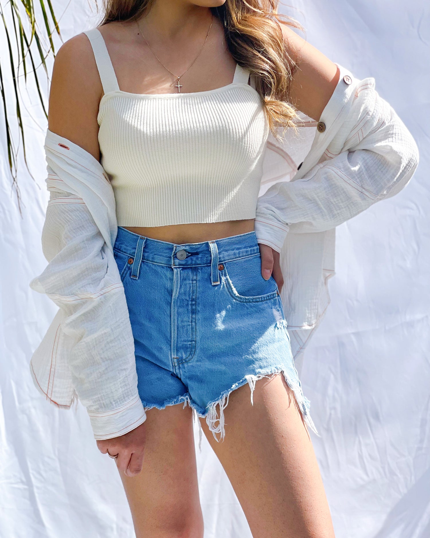A latina model is wearing an off-white ribbed knit crop top. It has a square neckline and wide shoulder straps. She is modeling it with denim shorts and a white pull-over oversized shirt. 